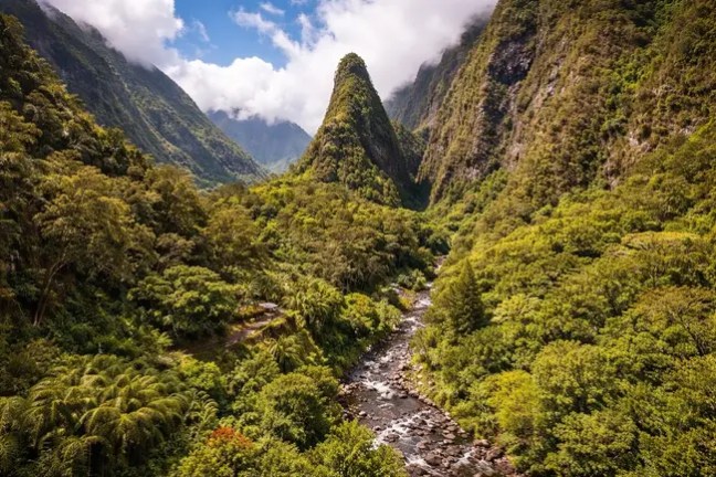 Lush green valley with a river, surrounded by steep hills and a peak under a cloudy sky.