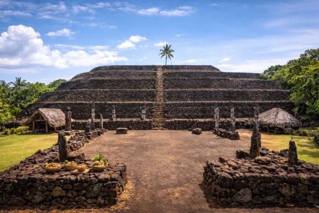 Ancient stone pyramid with palm tree and stone structures under blue sky.
