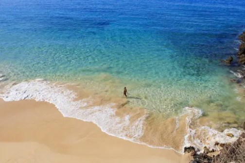 Person standing on a sandy beach with clear blue water nearby.