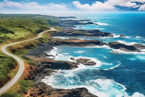 Coastal road winding along a rocky shoreline with blue ocean waves and green hills under a clear sky.