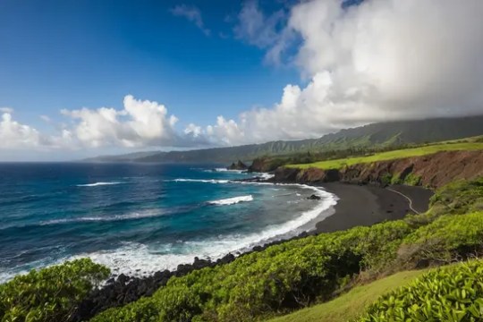 Coastline with black sand beach, lush greenery, and blue ocean under a cloudy sky.