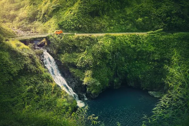 Orange car crossing bridge over waterfall and lush greenery.
