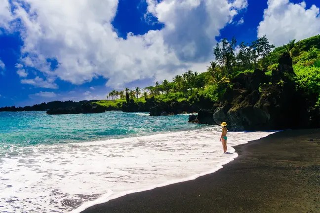 Person on black sand beach with bright blue sky and lush green trees.