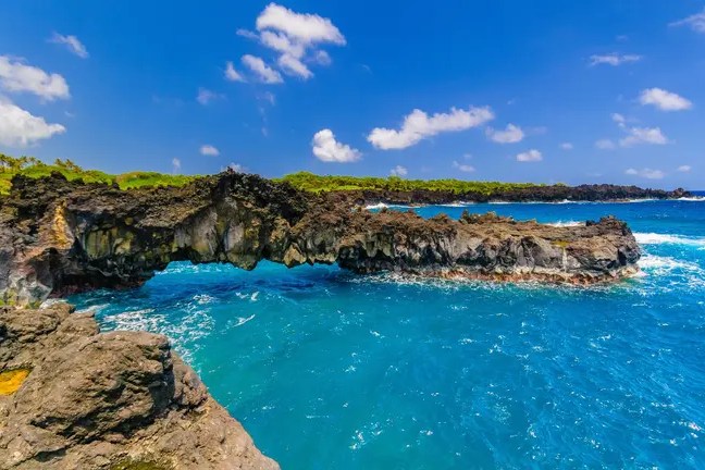 Rock arch over blue ocean with clear sky and scattered clouds.
