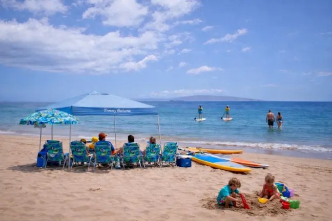 People relaxing under umbrellas on a sandy beach with others paddleboarding in the ocean.