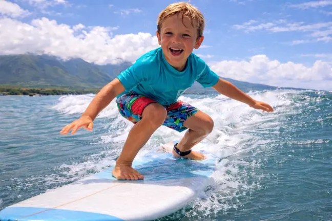 Smiling child surfing on a wave under a clear blue sky.